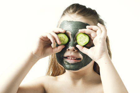 Funny little girl with a cosmetic mask of clay in a bath towel holding a piece of cucumber, like a mask, the concept of beauty and health, portrait indoors closeupの写真素材