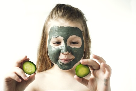 Funny little girl with a cosmetic mask of clay in a bath towel holding a piece of cucumber, like a mask, the concept of beauty and health, portrait indoors closeupの写真素材