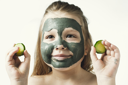 Funny little girl with a cosmetic mask of clay in a bath towel holding a piece of cucumber, like a mask, the concept of beauty and health, portrait indoors closeupの写真素材