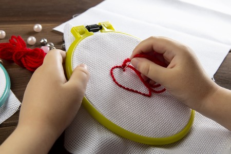 A little girl embroiders a heart on a white cloth view from above accessories for embroidery on wooden backgroundの写真素材