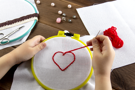 A little girl embroiders a heart on a white cloth view from above accessories for embroidery on wooden backgroundの写真素材
