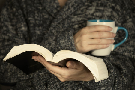 A young woman in a gray knitted sweater is reading a book.
