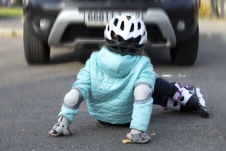 A little girl in a blue jacket and helmet on rollers fell in front of a moving car on a city street.の写真素材