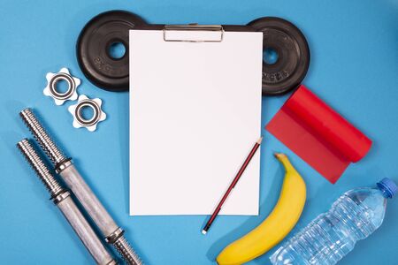 sports equipment for sports and fitness: barbell, dumbbells, elastic for fitness, jump rope, water in a bottle, elastic for fitness, top view, black background, in the middle a paper holder with empty white sheets and a pencilの写真素材