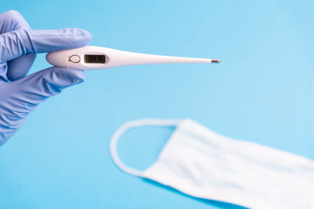 Doctor holds a digital thermometer on a blue background with a medical mask, closeup. Medical...