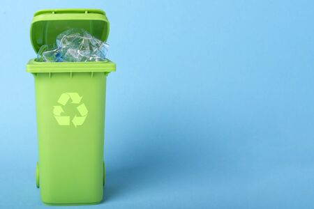 Green trash can with plastic waste and recycling icon on a blue background with place for text.の写真素材