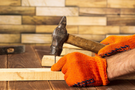 Close-up shot of a carpenter in orange work gloves hammers a nail into a wooden board.の写真素材