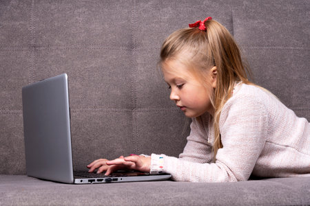 Little caucasian girl is engaged on a laptop while lying on a gray sofaの写真素材