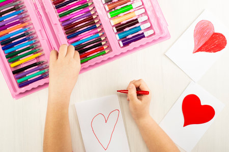 Little girl prepares cards with hearts for Valentine's Day. Drawing and applique work is done by the child using colored pencils or felt-tip pens. Children's drawing. View from above.の写真素材