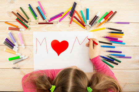 Little girl makes a greeting card to mom with a heart for mother's day. Drawing and applique work are done by the child using colored pencils or felt-tip pens. Happy family. Children's drawing. View from above.の写真素材