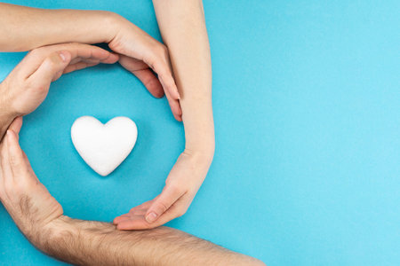 Hands of parents and a child surround a white heart on a blue background. Family concept, flat lay, top view.の写真素材