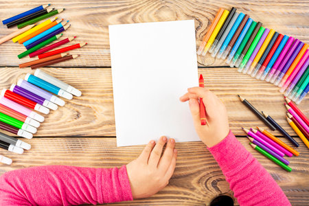 Little girl prepares to paint on a blank sheet of paper. Drawing is done by a child with colored pencils or felt-tip pens. Children's drawing. View from above.の写真素材