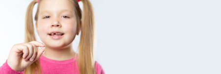 Little caucasian girl with long blond hair in pink clothes holds a pulled out milk tooth in her hands on a light background. Dental hygiene conceptの写真素材