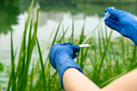 Gloved hands hold a chemical pipette and a glass slide with a drop of water. Sampling from open...