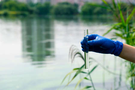 A gloved hand takes water into a test tube from a city reservoir. Urban waste water. Sampling from open water. Scientist or biologist takes a sample of water into a test tube.の写真素材