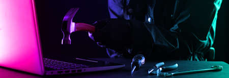 Criminal hacker in a hood in black clothes and balaclava destroys a laptop with a hammer on a black background.の写真素材