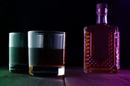 Two glasses with alcohol and a bottle on a wooden table in a dark room.の写真素材