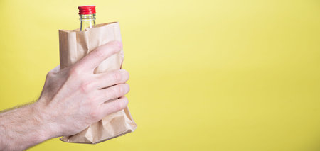 Male hand with a bottle of alcohol in a paper bag on a yellow background. Close up view with copy...