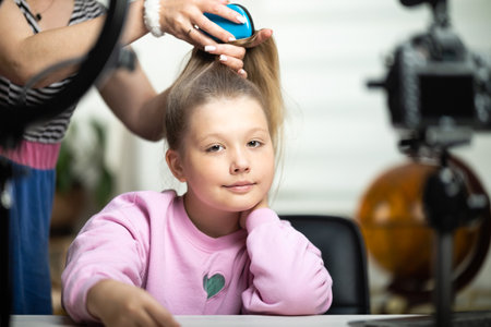 A beautiful blond teenage girl does her hair during a live broadcast. The video blogger is broadcasting live. Ring lamp and camera during the vloggers live broadcastの写真素材