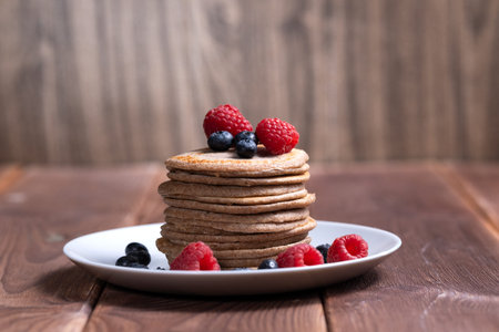 A stack of pancakes with fresh raspberries and blueberries on a light plate on a wooden backgroundの写真素材