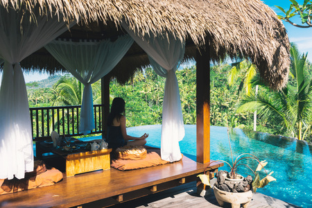 Girl meditating by the pool on a background of palms, Bali, Indonesia.The girl sits in a gazebo at a bali in a lotus pose and meditates with a view of the palms and the pool. Breakfast in Baliの写真素材