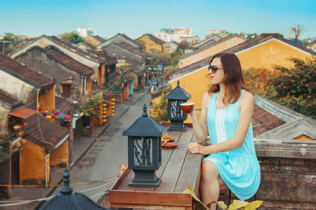 Tourist girl drinks coffee overlooking the old city of Hoi An, Vietnam. A young woman is enjoying the top view of the old Vietnamese city from the roof. A popular tourist destination in Vietnamの写真素材