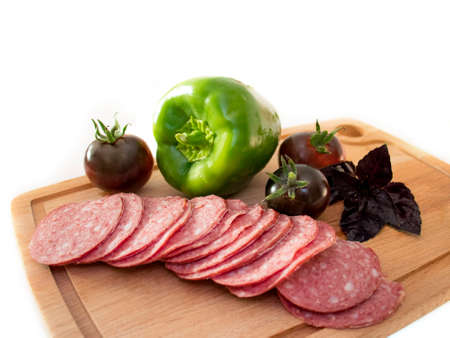 sausage slicing and fresh vegetables in the form of dark tomatoes, green paprika and fragrant purple basil on a wooden cutting doske. selective focus. isolate on whiteの写真素材