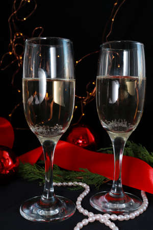 Two glasses of champagne on the festive table on a dark background. next to Christmas decor red balls, ribbon, bright garland in the background. Christmas composition. Selective focus.の写真素材