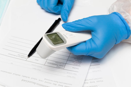 The doctor's hands in gloves hold a contactless thermometer to measure temperature before being...
