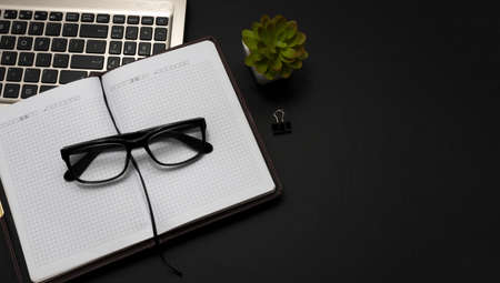 Top view of a stylish office desk with a plant an open notebook, glasses and a laptop. Study. Dark stylish workplace with laptop computer and glasses on black desk background, top view with copy spaceの写真素材