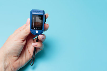 A female hand holds a pulse oximeter against a blue medical background with copy space. Banner. The...