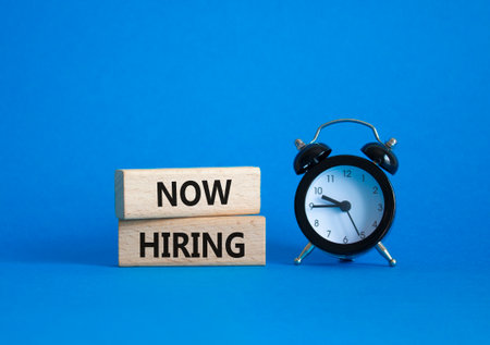 Now Hiring symbol. Wooden blocks with words Now Hiring. Beautiful blue background with alarm clock. Business and Now Hiring concept. Copy space.の写真素材