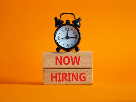Now Hiring symbol. Wooden blocks with words Now Hiring. Beautiful orange background with alarm clock. Business and Now Hiring concept. Copy space.の写真素材
