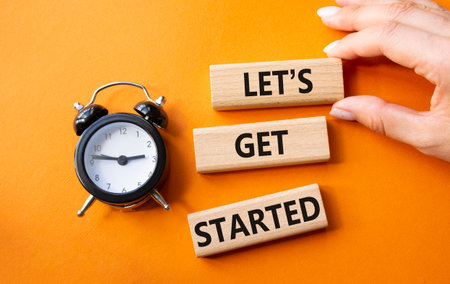Lets get started symbol. Concept words Lets get started on wooden blocks. Businessman hand. Beautiful orange background with alarm clock. Business and Lets get started concept. Copy space.の写真素材