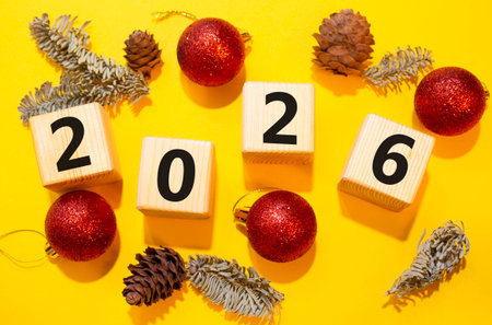 2026 New Year symbol. Wooden cubes with words 2026. Beautiful yellow background with Christmas balls and pine cones. Copy space. Business and 2026 happy new year conceptの写真素材