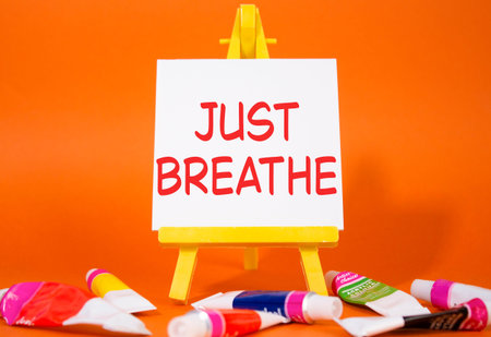 Just Breathe symbol. Wooden blocks with words Just Breathe. Beautiful orange background with paints and easel. Just Breathe concept. Copy space.の写真素材