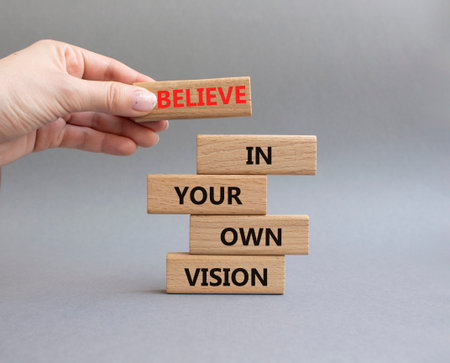 Wooden blocks with the words 'Believe in Your Own Vision'. A hand is holding a block with the word...