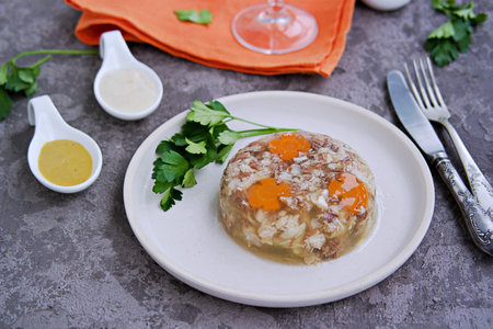 Cold appetizer, portioned jelly of pork, beef and chicken on a white clay plate on a gray concrete...