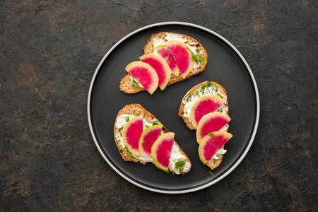 Light breakfast or snack, open sandwiches on whole grain bread with cream cheese, dill and watermelon radish slices on a black plate against a dark concrete background. Sandwich recipes. Healthy foodの写真素材