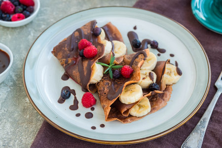 Thin chocolate pancakes stuffed with bananas, raspberries and blueberries, poured with chocolate sauce, on a gray ceramic plate on a light concrete background. Pancake recipes. Pancake dayの写真素材
