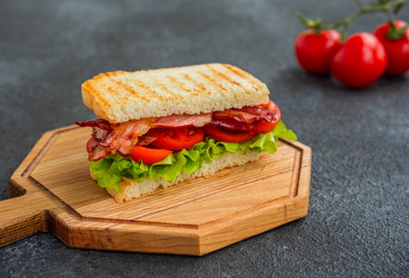 BLT sandwich with fried bacon, tomato and lettuce on a wooden board on a dark concrete background....