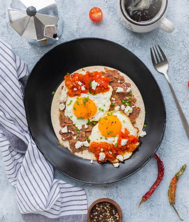 Huevos rancheros, Mexican fried egg on a wheat tortilla with tomato salsa, bean paste and feta cheese on a black plate on a light concrete background. Mexican food.の写真素材