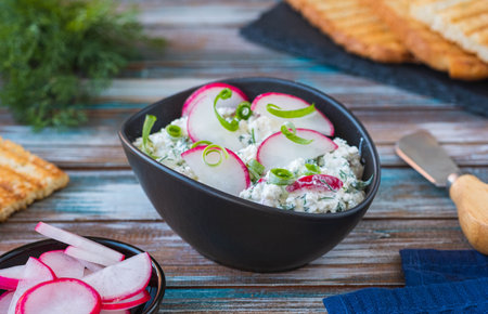 Appetizer, salted cottage cheese with dill, green onion and radish in a black bowl on a wooden background. Recipes with cottage cheese. Served with toasted white breadの写真素材