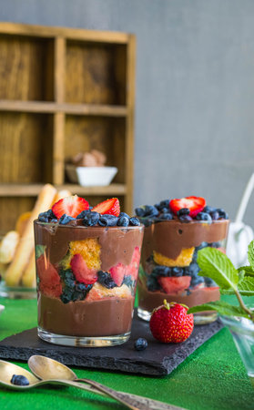 Summer dessert in layers in a glass with fresh strawberries, honeysuckle, biscuits and chocolate custard on a green concrete background. Desserts without baking, portioned desserts in a glass.の写真素材