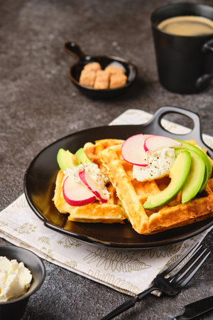Unsweetened cheese waffles with cream cheese, avocado and radish slices on a black plate on a dark concrete background. Breakfasts, savory waffles with toppings.の写真素材