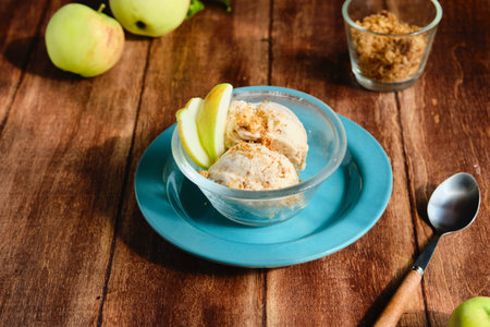 Two scoops of vanilla ice cream with shortbread crumble and caramelized apple slices in a transparent bowl on a brown wooden background. Apple desserts, ice cream.の写真素材