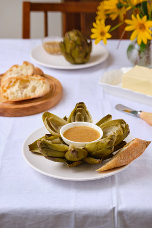 Steamed artichoke with vinaigrette sauce based on oil and mustard on a white plate on a light background. Served with fresh baguette and butter. French cuisine, delicacyの写真素材