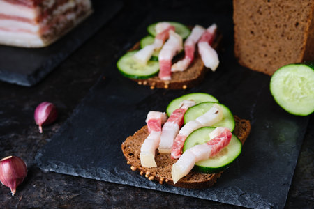 Two sandwiches with salted lard strips with meat strips and fresh cucumber on rye bread on a slate board on a black concrete background. Recipes for sandwiches, snacks. Ukrainian foodの写真素材