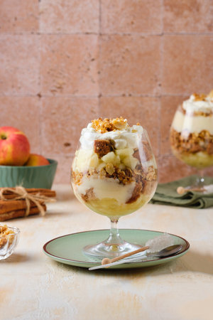Dessert, portioned apple trifle with oat crumble, spiced cake, custard and whipped cream in a glass goblet on a light concrete background. Apple desserts, portioned desserts in glassesの写真素材