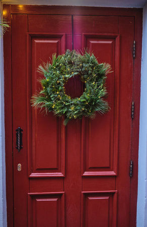Christmas wreath made of fir branches and garland hanging on red door outside. Decoration, holiday preparation, advent. Merry Christmasの写真素材